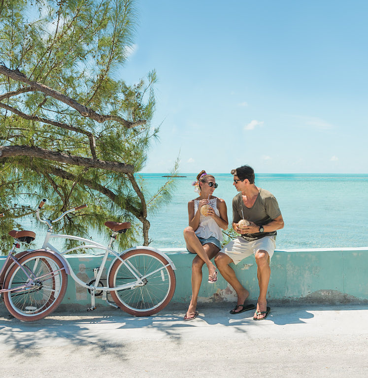 man and woman drinking from coconuts on the side of the road next to a bicycle with the ocean in the background
